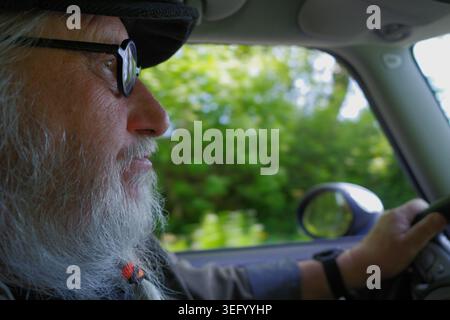 Homme senior avec une longue barbe blanche et des lunettes conduit une voiture. Il porte un chapeau et semble concentré sur la route. L'arrière-plan à l'extérieur du véhicule sh Banque D'Images