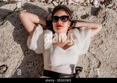 Mode femme méditerranéenne couchée sur des rochers ensoleillés en robe blanche et lunettes de soleil pendant les vacances d'été sereines Banque D'Images