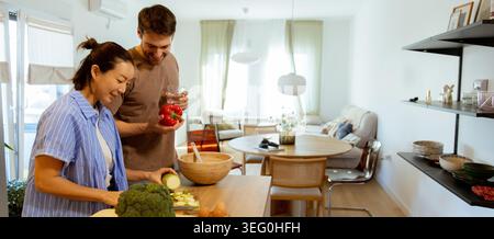 Une coréenne et un homme caucasien cuisinent dans une cuisine moderne. La femme prépare des légumes pendant que l'homme tient un verre d'eau et le regarde Banque D'Images