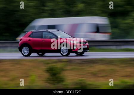 Une photographie panoramique nette d'un crossover rouge Kia Stonic conduisant sur une route mouillée, avec un bus de passagers blanc et rouge se déplaçant rapidement dans le creat d'arrière-plan Banque D'Images