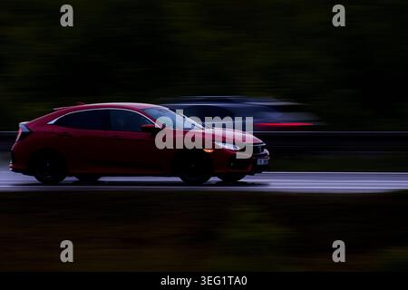 Photo panoramique dynamique d'une berline moderne rouge à hayon roulant rapidement sur une autoroute mouillée lors d'une journée sombre et sombre avec un arrière-plan vert foncé flou Banque D'Images