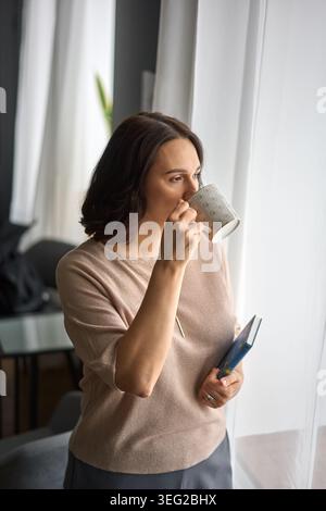 Une femme dans un pull neutre prend une gorgée de café tout en regardant par une fenêtre lumineuse, tenant un petit livre. La scène capture un moment privé des hommes Banque D'Images