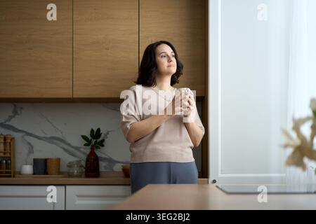 Une femme sereine tient une tasse en céramique avec les deux mains, regardant par une fenêtre dans une cuisine moderne. La scène capture un moment de pleine conscience tranquille et emo Banque D'Images