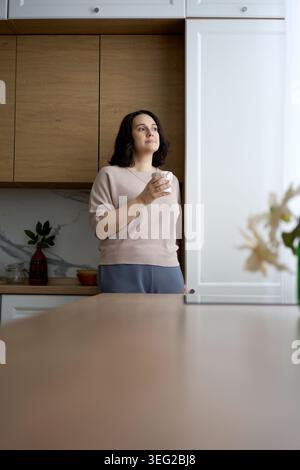 Debout dans une cuisine lumineuse, une femme regarde soigneusement au loin tout en tenant une tasse. L'éclairage haut de gamme et le décor minimaliste soulignent un Banque D'Images
