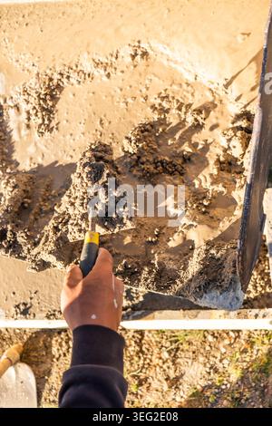Gros plan d'un ouvrier de la construction utilisant une truelle pour lisser et niveler le béton frais dans une tranchée de coffrage extérieur, créant un bord de fondation propre o Banque D'Images