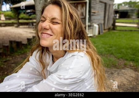 Portrait en gros plan d'une femme souriant les yeux fermés à l'extérieur, créant un moment de vie naturel, ludique et authentique à la campagne. Banque D'Images