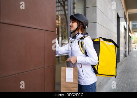 Femme souriante livreur de nourriture portant un casque et un sac à dos isolé, livrant la commande à emporter à l'entrée d'un bâtiment avec un sac en papier à la main, urba Banque D'Images