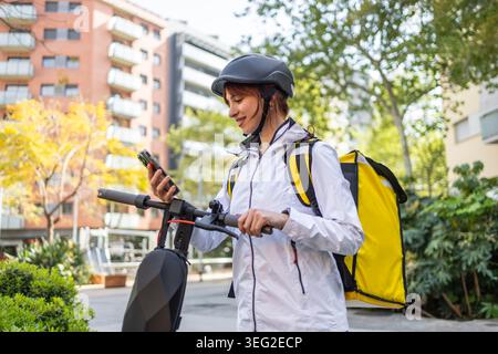 Femme souriante coureur de livraison de nourriture dans le casque vérifie son smartphone à côté d'un scooter électrique, portant un sac à dos isolé jaune pour couri urbain rapide Banque D'Images