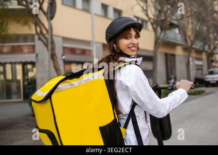 Femme souriante coureur de livraison de nourriture portant un casque et portant un sac à dos isolé jaune tout en montant un e-scooter dans une rue moderne de la ville, prêt pour Banque D'Images