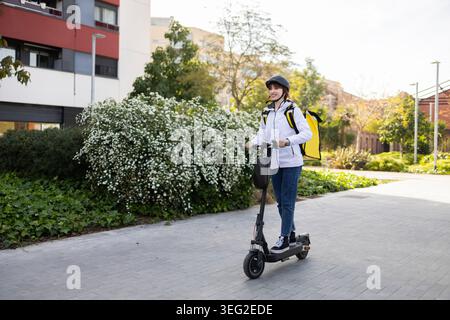 Femme souriante livreur de nourriture monte un scooter électrique le long d'un trottoir urbain ensoleillé, portant casque et sac à dos isolé, livrant des repas quic Banque D'Images