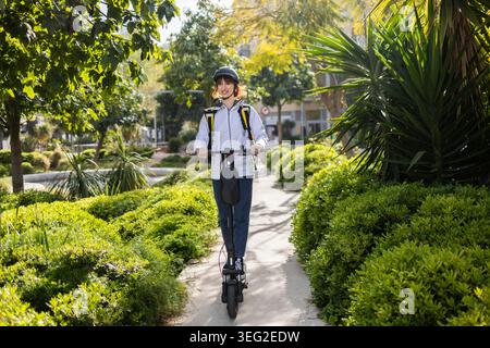 Femme souriante coureur de livraison de nourriture portant un casque et un sac à dos monte un scooter électrique le long d'un chemin de parc ensoleillé, représentant des couri rapides et respectueux de l'environnement Banque D'Images