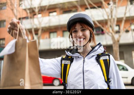 Femme souriante livreur portant un casque et une veste remet un sac en papier à emporter au client à l'extérieur dans une rue de la ville, mettant en vedette la livraison de nourriture rapide Banque D'Images