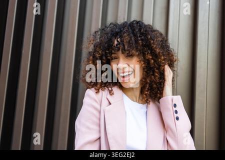 Femme hispanique aux cheveux bouclés souriant joyeusement à l'extérieur, habillée d'un blazer rose élégant, incarne un style de vie moderne et une énergie positive, debout a Banque D'Images