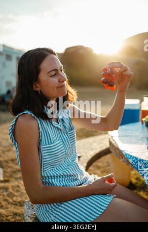 Jeune femme en robe rayée profitant d'un pique-nique d'été à l'extérieur, tenant un bouquet de raisins à une table pendant l'heure d'or, ambiance détendue de vacances à natu Banque D'Images