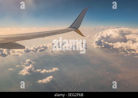 Vue depuis une fenêtre d'avion de passagers montrant l'aile s'étendant sur des nuages moelleux à l'heure dorée, avec la lumière douce du soleil, le ciel bleu et les landsca lointains Banque D'Images