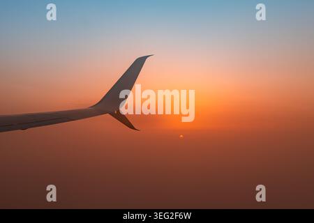 Silhouette d'une aile d'avion coupant à travers des nuages brumeux lors d'un coucher de soleil orange vibrant, vue depuis une fenêtre passager, avec une lumière douce et calme tr Banque D'Images