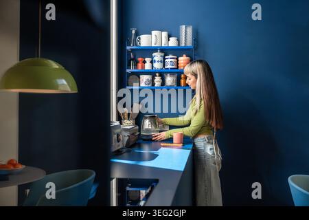 Vue de côté d'une jeune femme dans un tablier préparant le thé avec une bouilloire électrique à un comptoir de cuisine à domicile, avec mur bleu, étagère ouverte, tasses et daylig chaud Banque D'Images