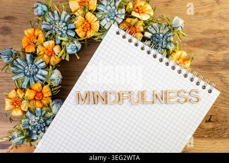 Mot de pleine conscience écrit avec des lettres en bois sur un cahier et une couronne de fleurs colorées, Banque D'Images