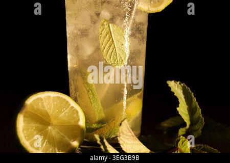 Cocktail Mojito dans un grand verre présentant des bulles gazeuses rafraîchissantes, des quartiers de citron vert, des feuilles de menthe fraîche et de la glace, accompagné d'un citron vert coupé en deux sur un d. Banque D'Images