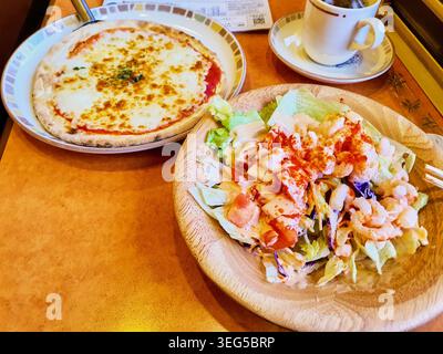 Une mise en place de table de restaurant comprenant une pizza Margherita sur une assiette en céramique décorative accompagnée d'une salade de crevettes fraîches servie sur une assiette en bois. Le Banque D'Images