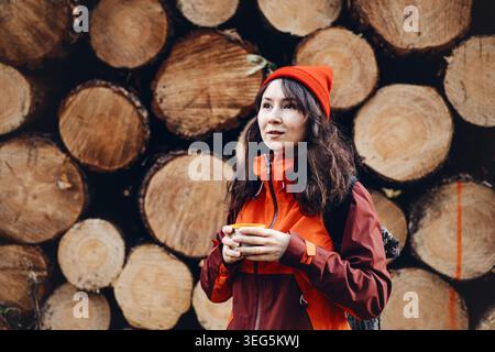 La femme se tient devant des bûches empilées tenant la tasse. Moments de voyage en solo, prendre un souffle en plein air, pause boisson chaude dans la nature. Banque D'Images