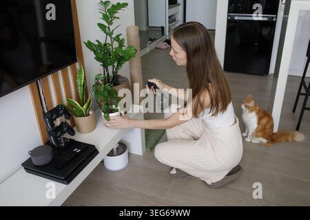 Femme brune avec chat animal de compagnie arrosant plante d'intérieur à la maison, passe-temps de soin des plantes, vie domestique, jardinage Banque D'Images
