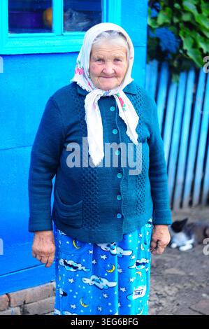 Une femme âgée souriante portant un cardigan tricoté et un foulard floral pose dehors devant un mur bleu vif et une clôture Banque D'Images