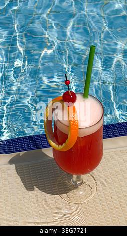 Cocktail tropical rafraîchissant avec tranche d'orange et cerise servi à côté d'une piscine, eau bleue vibrante reflétant la lumière du soleil en arrière-plan Banque D'Images