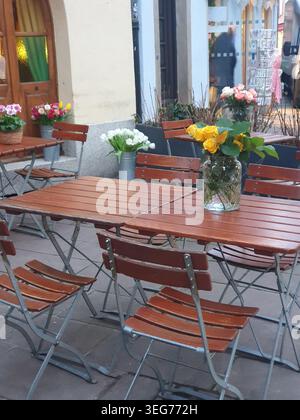 Des arrangements floraux lumineux sur des tables de bistrot en bois créent une atmosphère chaleureuse de café en plein air dans une charmante cour, avec des roses, des tulipes et du pot p Banque D'Images