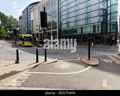 Manchester, Royaume-Uni - 01 mai 2026 : intersection urbaine à Manchester Royaume-Uni avec un bus jaune, des pistes cyclables, des bâtiments en verre et des piétons. Banque D'Images