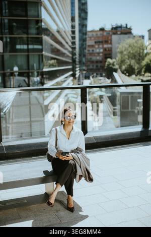 Une femme est assise sur une marche dans une ville pendant la journée. Elle est habillée d'une chemise blanche et d'un pantalon noir. Elle regarde son téléphone tout en profitant de la chaude w Banque D'Images