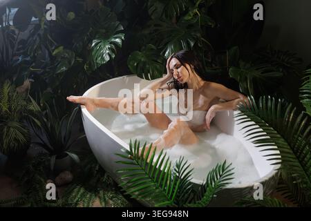 Une scène sereine avec une femme profitant d'un bain moussant dans une baignoire moderne, entourée d'un feuillage tropical luxuriant, parfait pour les promotions spa et bien-être. Banque D'Images