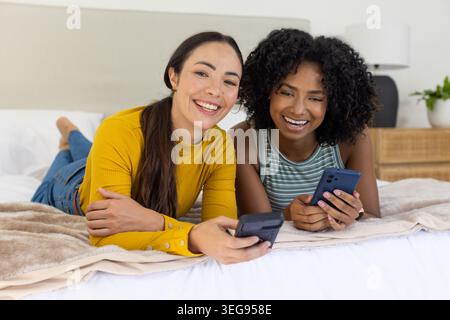 Diverses amies féminines allongées sur le ventre sur le lit avec une couette blanche tenant des smartphones foncés et bleus Banque D'Images