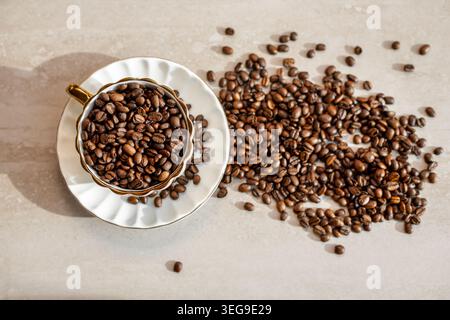 Grains de café torréfiés renversés à côté de la tasse. Les grains de café torréfiés remplissent une tasse élégante et se répandent sur une surface neutre, créant un café chaleureux inspiré du café Banque D'Images