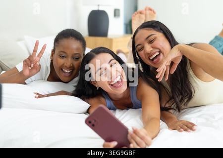 Diverses amies féminines riant et prenant selfie sur le lit à la maison avec smartphone rose Banque D'Images