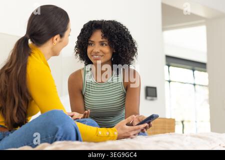 Diverses amies féminines parlant sur le lit dans une chambre lumineuse, l'une portant le dessus moutarde tenant le smartphone Banque D'Images