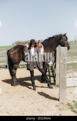 Une femme pose près d'un cheval noir dans un cadre de ranch. Elle porte un chapeau et un manteau tout en se tenant debout sur un sol sablonneux à côté du cheval. Le soleil brille en un cle Banque D'Images