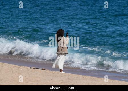 Jeune femme debout sur une plage de sable face à l'appareil photo, tenant un smartphone, semblant prendre une photo ou une vidéo de l'océan. Banque D'Images