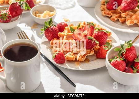 Gaufres belges avec fraises fraîches et miel sur plaque blanche sur fond de nappe blanche. Petit déjeuner d'été sucré dans la lumière du matin ensoleillée et lumineuse Banque D'Images