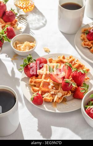 Gaufres belges avec fraises fraîches et miel sur plaque blanche sur fond de nappe blanche. Petit déjeuner d'été sucré dans la lumière du matin ensoleillée et lumineuse Banque D'Images