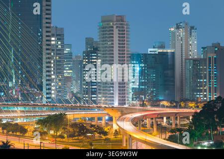 La longue exposition capture les traînées de lumière sur les rampes d'autoroute courbes menant à un pont avec l'horizon dense de Sao Paulo Brésil Banque D'Images