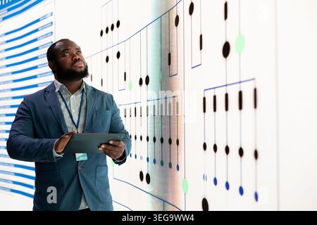Inspection des plans de circuits imprimés, écran et technicien analysant la topologie du réseau. L'homme afro-américain dans l'usine de micropuce examine la disposition de fabrication de circuits électriques sur le mur numérique, validant la disposition Banque D'Images
