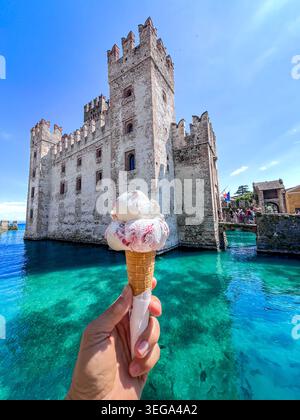 Une main tenant un délicieux cornet de crème glacée à double cuillère devant le château historique de Scaligero entouré par les eaux turquoises du lac de Garde en S. Banque D'Images