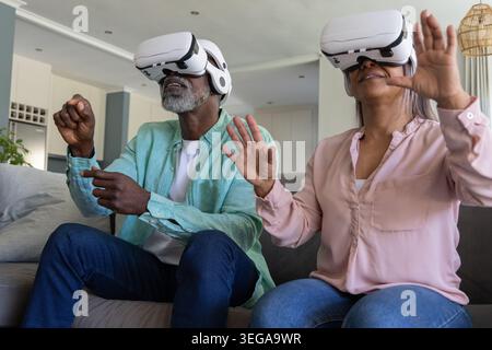 Couple mature utilisant des casques blancs VR, homme afro-américain et femme faisant des gestes sur le canapé Banque D'Images