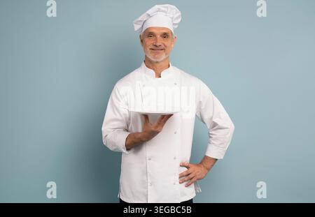 Portrait du chef, assiette vide et sourire, offre promotionnelle spéciale de menu ou placement de produit de restaurant. Cuisinier heureux en uniforme isolé sur BA bleu Banque D'Images