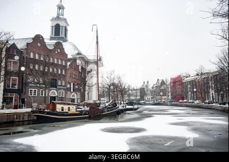 Une scène hivernale sereine à Amsterdam avec des bâtiments traditionnels en briques recouverts de neige et un canal gelé avec des bateaux amarrés le long des côtés. Banque D'Images