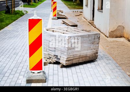 Une palette enveloppée de pavés et de barrières de sécurité rouges et jaunes repose sur un trottoir gris partiellement fini lors d'un pro de construction de rues urbaines Banque D'Images