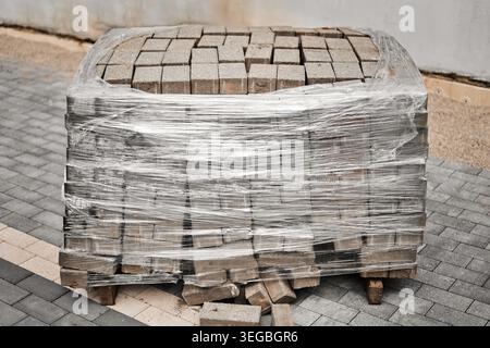 Une palette de pavés en béton gris enveloppé dans un film plastique transparent, assis sur un pavé fini prêt pour une construction extérieure ou l'aménagement paysager Banque D'Images