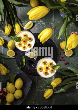 Tartes rafraîchissantes à la crème de citron, garnies de graines de grenade et de notes d'agrumes, sur des carreaux vert foncé avec des tulipes jaune vif et des citrons frais pour un Banque D'Images