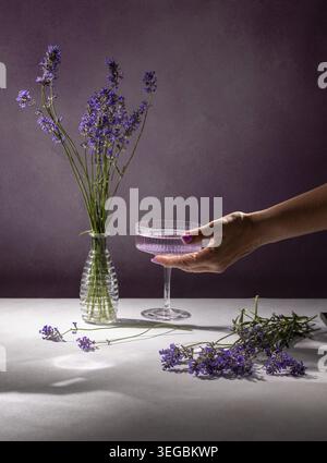 Tenant un cocktail de lavande avec des fleurs de lavande fraîches dans un vase, étalé sur une table blanche sur un fond violet, créant un arôme détendu Banque D'Images
